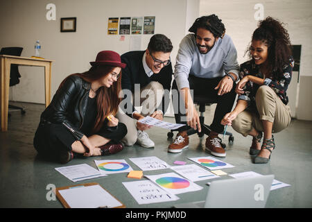 Cheerful business colleagues sitting on floor in office and discussing work. Teammates with business papers and laptop spread on the floor discussing Stock Photo