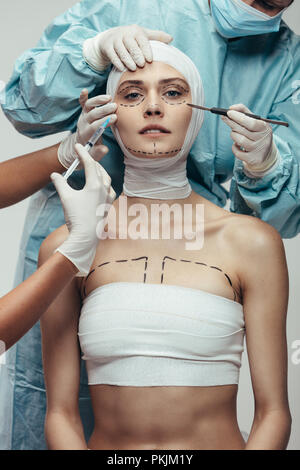 Portrait of young woman wrapped in medical bandages while beautician doctors with syringe and scalpel near her face. Female having a face lift surgery Stock Photo