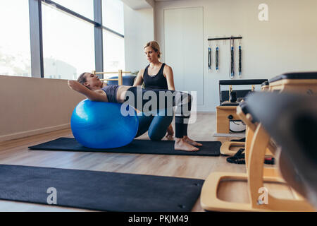 Woman doing pilates workout at the gym using an exercise ball while her trainer looks on. Pilates woman lying with her back on a fitness ball. Stock Photo