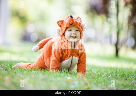 Cute smiling baby girl dressed in fox costume crawling on lawn in park Stock Photo