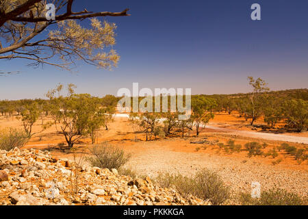 View from stony hill of Australian outback landscape with road slicing across plains, dotted with trees, that stretch to horizon under blue sky in Qld Stock Photo