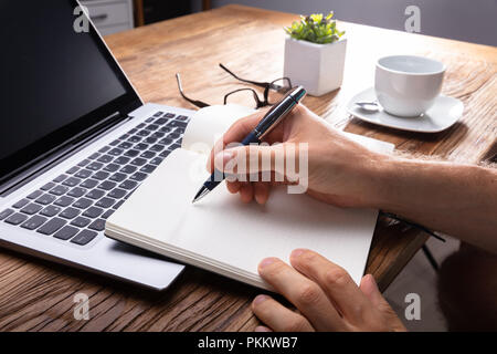Person's Hand Writing On Black Notebook Using Pen Over Laptop Stock Photo