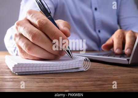 Person's Hand Writing On Black Notebook Using Pen Over Laptop Stock Photo