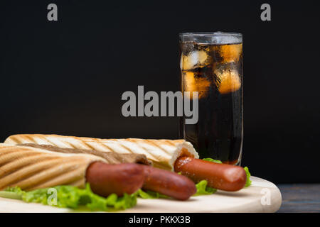 Tall glass of soda with ice next to plate of hot dogs wrapped in bread on top of large leaves of lettuce Stock Photo
