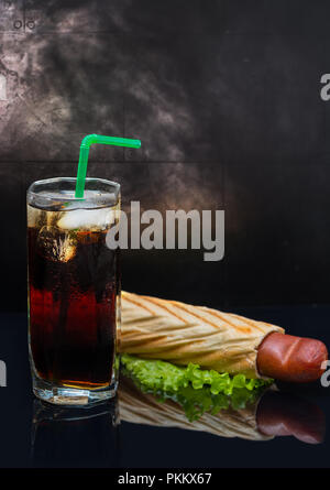 Soda with ice and pigs in a blanket hot dog on green lettuce over dark reflective background. Steam in background. Stock Photo