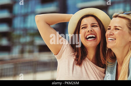 Two laughing young female friends standing arm in arm having a good time together in the city in summer Stock Photo