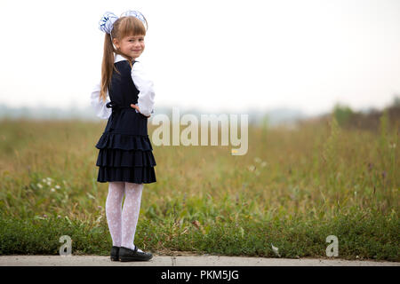 Full-length portrait of cute adorable serious thoughtful first grader girl in school uniform and white bows in long blond hair on blurred light green  Stock Photo