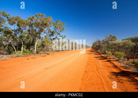 Long red Australian outback road slicing through landscape cloaked with bushland and tall gumj trees & stretching to horizon under blue sky Stock Photo