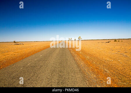 Narrow Australian outback road slicing through arid landscape devoid of vegetation during drought & stretching to distant horizon under blue sky Stock Photo