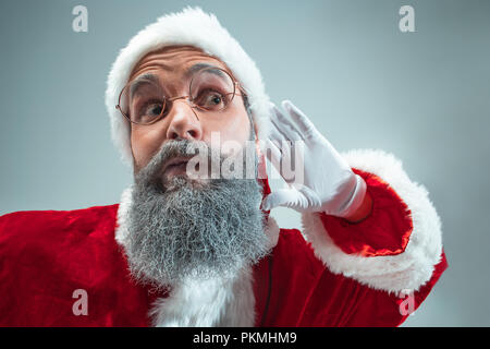 Funny eavesdropping guy with christmas hat posing at studio. New Year Holiday. Christmas, x-mas, winter, gifts concept. Man wearing Santa Claus costume on gray. Copy space. Winter sales. Stock Photo