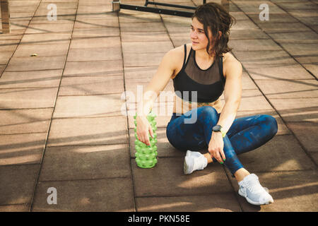 theme sport and rehabilitation sports medicine. Beautiful strong slender Caucasian woman athlete sits next foam roller green field street workout to r Stock Photo