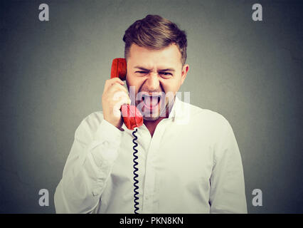 Young man in white shirt holding red telephone handset and screaming in frustration on gray background. Stock Photo