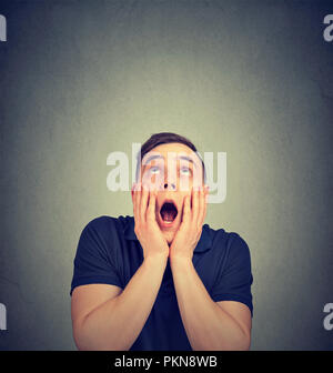 Casual young man holding hands on cheeks and looking up in expression of fear and amazement Stock Photo