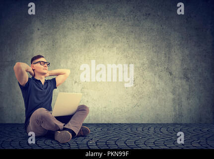 Young casual man in glasses holding hands behind head while sitting with laptop on gray background and relaxing Stock Photo