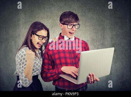 Young man and woman in eyeglasses looking super happy while watching laptop on gray background Stock Photo