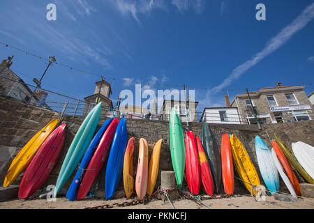 Colourful kayaks, canoes and surfboards standing upright on the sandy beach of Mousehole in Cornwall, UK with the quaint fishing village behind. Stock Photo