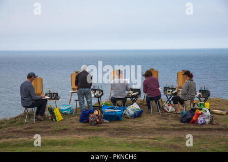 An art teacher teaching a group of mature students how to paint whilst sitting at an easel overlooking a beautiful ocean landscape scene Stock Photo