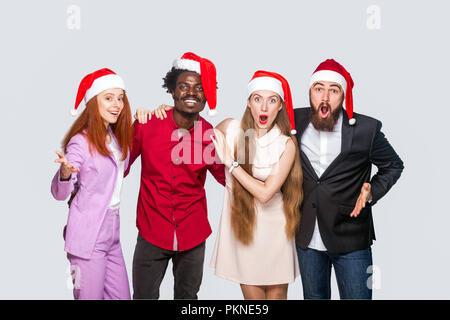 Group of beautiful happy handsome friends in red cap standing and celebrating new year and looking at camera with amazed surprised face. indoor studio Stock Photo