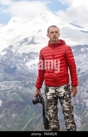Male photographer with camera in hands stands on a background of mountains Elbrus. Ice the top of mount Elbrus behind men Stock Photo