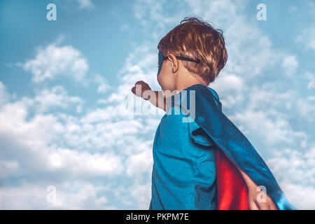 Little boy wearing costume of a superhero with  raised up one hand over sky background, preparing to save the world, kid plays in superman Stock Photo