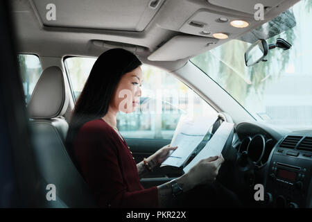 Chinese female manager working in car. Successful Asian businesswoman using laptop computer and looking at paperwork. Stock Photo