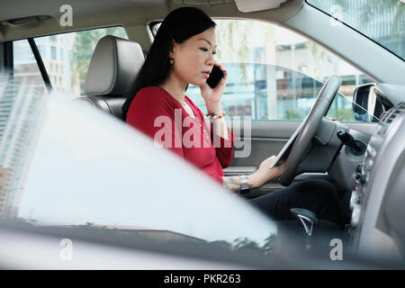 Efficient Chinese female manager working in car. Successful Asian businesswoman smiling and talking on cell phone. Stock Photo
