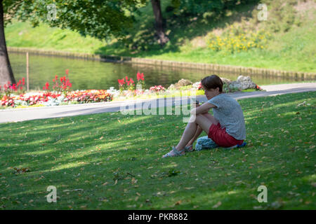 RIGA, LATVIA - JULY 18, 2018: A young man sits in a park on the ground and looks at the phone. Stock Photo