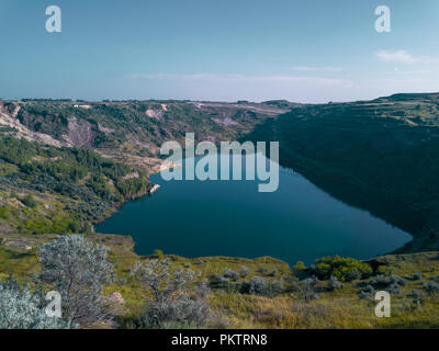 Aerial drone view of abandoned quarry with lake appeared on the bottom, the mirror surface of the water in the sunny evening, overgrown with forest sh Stock Photo