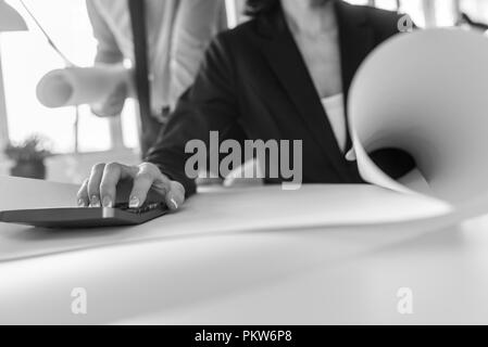 Greyscale image of businesswoman working on calculator as her partner stands behind her in a low angle view. Stock Photo
