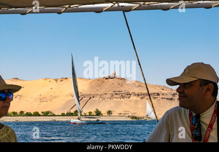 Egyptian man and woman in traditional felucca sailing boat with view of hilltop tomb, Qubbet el Hawa, River Nile, Aswan, Egypt, Africa Stock Photo