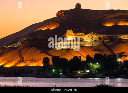 Night view across Nile river to hilltop Qubbet el Hawa tomb, Dome of the Winds, and ancient hillside tombs lit up, Aswan, Egypt, Africa Stock Photo