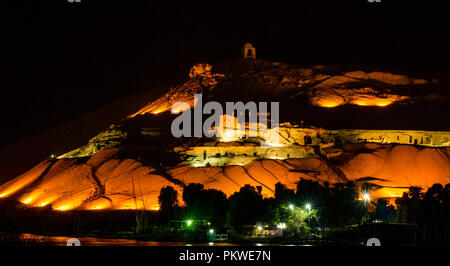 Night view across Nile river to hilltop Qubbet el Hawa tomb, Dome of the Winds, and ancient hillside tombs lit up, Aswan, Egypt, Africa Stock Photo