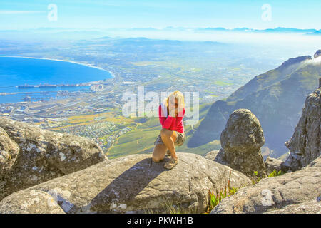 Caucasian traveler sitting on the rocks after hiking at Table Mountain. Sporty woman enjoying aerial views of Port of Cape Town from the top of Table Mountain NP in Western Cape, South Africa. Stock Photo