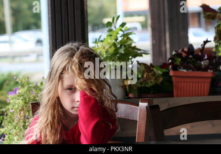 little girl sits alone in a cafe, covered half of her face with her hand and blond hair, a red jacket, flowers in the background, a chair next to her, Stock Photo