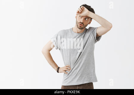 Phew so close. Portrait of relieved handsome dark-haired man with beard in grey t-shirt whiping sweat from forehead breathing out with folded lips and gazing left, solving hard problem Stock Photo