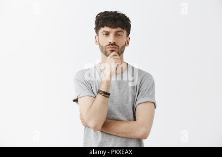 Waist-up shot of good-looking thoughtful male genius with dark hair and beard holdin hand on chin and pursing lips looking seriously at camera having plan or considering how make things right Stock Photo