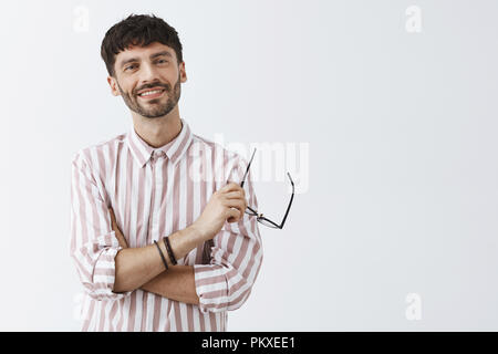 Cool charming bearded caucasian guy with dark short hairstyle crossing hands on chest holding glasses in hand smiling with carefree and confident expression relaxing after giving speech on lecture Stock Photo