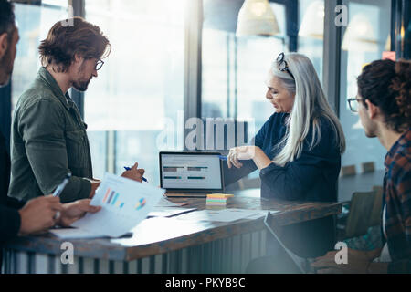 Senior business woman showing a presentation on laptop to colleagues around a table. Small presentation during new project discussion. Stock Photo