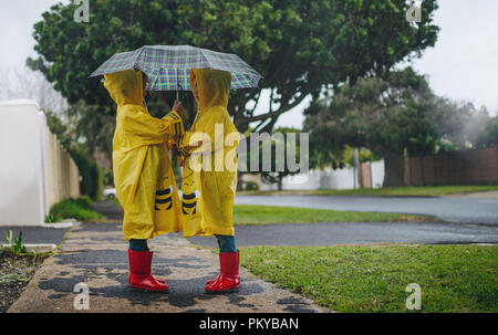Two little girls wearing waterproof coats and rubber boots standing under the umbrella outdoors. Little sisters outdoors on a rainy day. Stock Photo