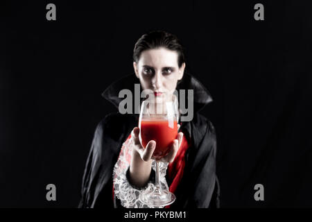 Woman dressed up as a vampire for halloween holding glass of red drink. Studio shot in dramatic lights of a young female in dracula costume on black b Stock Photo