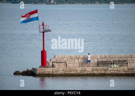 Croatian flag flying in wind on lighthouse in harbor, daytime, stone pier, man, facing away looking over stone wall Stock Photo