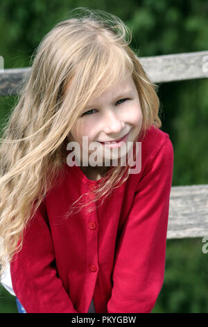 little blonde girl with curly long hair, portrait, in a red sweater in the sun, vertical photo, looks at the camera and smiles, in the background Stock Photo