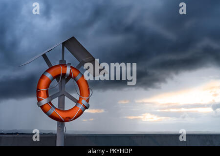 Orange lifebuoy on a stand on harbor pier against dramatic clouds in the sky. Stock Photo