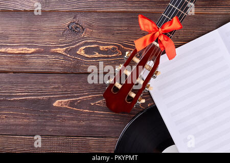 Guitar and music sheets on wooden background. Guitar handle with red bow and gramophone record with copy space. Old and modern musical equipment. Stock Photo