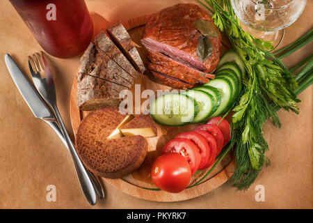 Simple lunch theme still life. Cheese, ham, vegetables and greenery on round wooden cutting board with wine bottle, glass and tableware top view Stock Photo