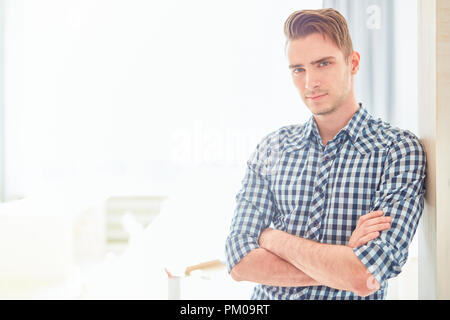 Serious guy with his arms crossed on chest looking at you while leaning against wall of modern design studio Stock Photo