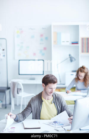 Young economist comparing financial data on papers while sitting by workplace in office Stock Photo
