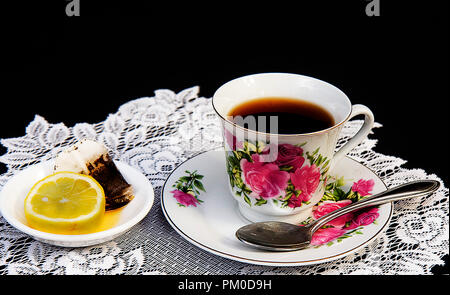 Old fashion cup of tea on lace doily with slice of lemon and tea bag to the side.  Isolated on black background. Stock Photo