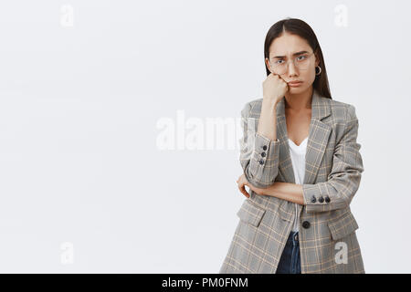 Portrait of moody sad girlfriend in fashionable outfit and glasses, leaning head on fist, sulking and frowning, expressing regret and disappointment, standing upset over gray background Stock Photo