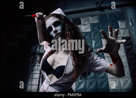 Horror shot: the horrible wicked mad nurse (doctor) in bloody uniform killing by syringe. Zombie woman (living dead). Monster from nightmare Stock Photo